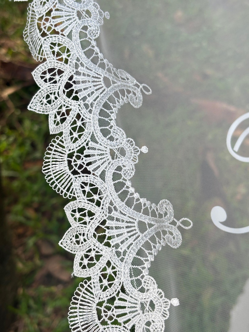 May include: A white bridal veil featuring an intricate scalloped lace edge with detailed floral and fan-like embroidery patterns. The sheer, lightweight tulle fabric extends from the decorative lace border, showcasing a delicate and fine mesh texture.
