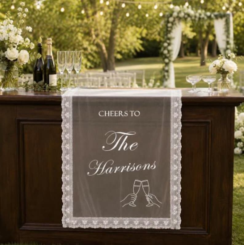 May include: A white sheer table runner with lace trim featuring the printed text CHEERS TO The Harrisons and a line art graphic of two hands holding champagne flutes. The runner is draped over a dark wooden bar surface in an outdoor setting.
