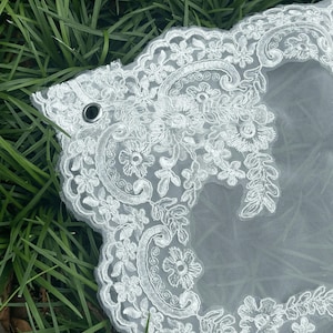 May include: A white wedding veil featuring intricate floral embroidery along the scalloped edges. The sheer tulle fabric is accented with detailed thread work in a botanical pattern, and a small metal grommet is visible on the upper left corner.