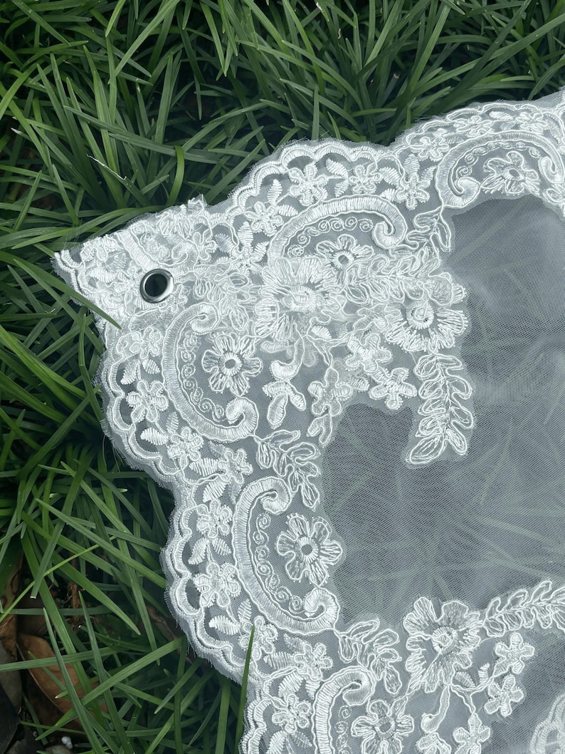 May include: A white wedding veil featuring intricate floral embroidery along the scalloped edges. The sheer tulle fabric is accented with detailed thread work in a botanical pattern, and a small metal grommet is visible on the upper left corner.