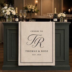 May include: A beige fabric table runner featuring a black border and centered black text that reads CHEERS TO, the initials TR in a large script font, THOMAS & ROSE, and EST. 2026. The runner hangs over the front of a dark gray wooden bar.