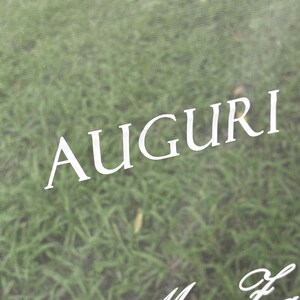 May include: A transparent acrylic sign featuring the white text AUGURI in a clean, sans-serif font, positioned above a partial line of white cursive script text that begins with the words Mrs Fa.