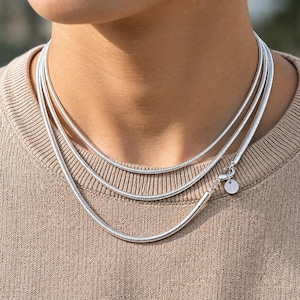 May include: A multi-strand silver snake chain necklace featuring a smooth, flexible metallic texture. The necklace consists of three layered strands of equal thickness, secured with a lobster claw clasp and a small, round, blank silver charm.