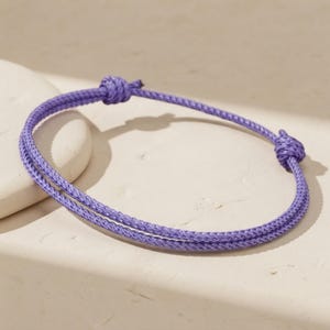 May include: A purple cord bracelet featuring a double-strand design. The bracelet is constructed from a thin, woven synthetic material and includes two small, knotted sliding closures that allow for adjustment of the circumference.