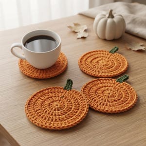 Könnte beinhalten: Drei runde, orangefarbene, gehäkelte Untersetzer mit kleinen grünen Stielen, die wie Kürbisse gestaltet sind. Ein Untersetzer trägt eine weiße Keramiktasse mit schwarzem Kaffee, während die anderen beiden in der Nähe auf einer Holzoberfläche liegen.