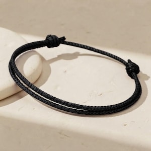 May include: A black cord bracelet featuring a double-strand design. The bracelet is constructed from thin, braided black string and includes two prominent, hand-tied knots that serve as adjustable closures for the circular wrist accessory.
