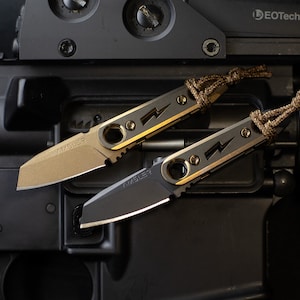 May include: Two small knives with a lightning bolt design and a lanyard are displayed. One knife has a gold blade and the other has a black blade. The knives are resting on a black surface with the word "SAFE" visible.