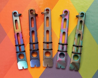 May include: Five multi-colored metal keychains with a rectangular design, each featuring a key ring and a small, slotted end. The keychains have a variety of colors, including blue, purple, green, and gold, and are accented with black rubber rings.