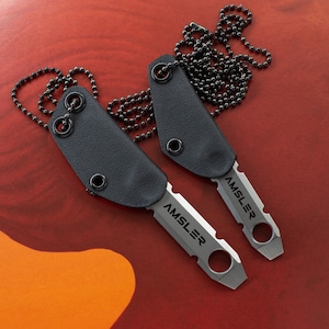 May include: Two small, silver knives with the word "AMSLER" engraved on the blades, each with a black leather sheath and a black ball chain necklace. The knives are displayed on a red surface.