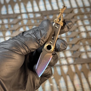 May include: A small, pocket-sized knife with a multi-colored blade. The knife has a dark gray handle with a lightning bolt design and a gold-colored lanyard. The blade has a rainbow-like finish.