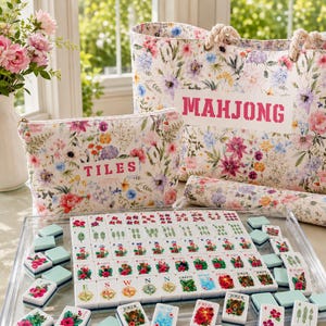 May include: A floral print tote bag with the text MAHJONG, a matching zippered pouch with the text TILES, and a set of white mahjong tiles featuring colorful botanical and animal illustrations arranged in a clear acrylic tray.