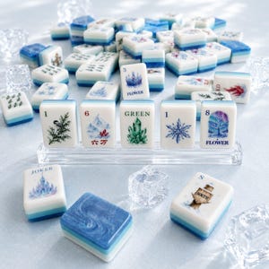 May include: A set of white and blue mahjong tiles featuring various illustrations including a bird, mountains, crystals, snowflakes, and trees. Some tiles display text such as 1, 3, 6, 8, GREEN, FLOWER, JOKER, and SOUTH. Several clear acrylic ice-like cubes are scattered among the tiles.
