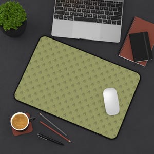 May include: A rectangular desk mat featuring a repeating olive green pattern of small, angled chevron shapes. The mat has a black border and is positioned on a dark surface alongside a laptop, a white computer mouse, notebooks, pens, and a coffee mug.