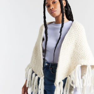 Puede incluir: Un chal triangular de punto grueso en color crema, con un patrón de tejido texturizado y largos flecos sueltos a lo largo de los bordes inferiores. El chal se lleva sobre una camiseta de manga larga color lila y unos vaqueros azul oscuro.