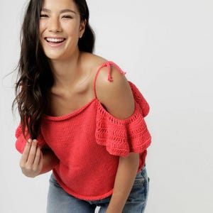 Puede incluir: Una blusa de punto en color rojo coral brillante con diseño de hombros descubiertos, tirantes finos y mangas cortas con volantes. La prenda presenta un escote recto sencillo y una textura de tejido a mano, combinada con pantalones vaqueros azul claro.