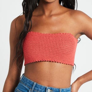 Puede incluir: Un top tipo tubo sin tirantes confeccionado con hilo de color coral en un patrón de punto de ganchillo denso. La prenda presenta un escote recto y un borde festoneado de ganchillo a juego en los dobladillos superior e inferior, con vaqueros azules.