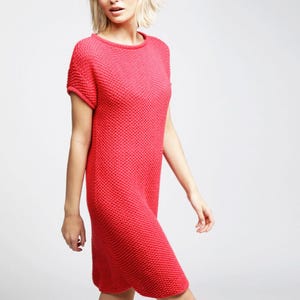 Puede incluir: Un vestido de punto rojo brillante con un patrón de tejido texturizado tipo panal, mangas cortas con puños enrollados y un cuello redondo sencillo. La prenda tiene una silueta recta y relajada que llega justo por encima de la rodilla.