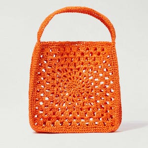 Puede incluir: Un bolso de mano de color naranja brillante elaborado con un patrón de ganchillo cuadrado que presenta un diseño calado. El bolso tiene un asa redondeada única unida a la parte superior, y toda la estructura está hecha de un material tejido texturizado.