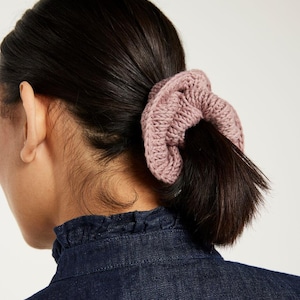 Puede incluir: Un coletero para el pelo de color rosa empolvado hecho de hilo grueso de punto. El accesorio presenta un diseño circular fruncido con una textura acanalada visible, colocado alrededor de una coleta de pelo oscuro para sujetarlo firmemente.