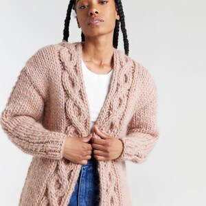 Puede incluir: Un cárdigan de punto grueso en color rosa empolvado que presenta un prominente patrón de trenzas verticales a lo largo de los paneles frontales y las mangas. La prenda tiene mangas largas, diseño abierto y una textura de hilo grueso.