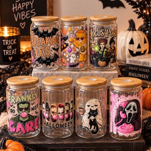 Może przedstawiać: Siedem przezroczystych szklanek z bambusowymi pokrywkami, każda z unikalnymi grafikami o tematyce Halloween, w tym nietoperzami, misiem, siostrami Sanderson, Frankensteinem, duchem z kotem oraz zamaskowaną postacią z różowym telefonem.