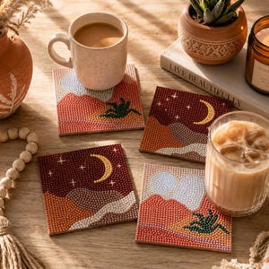 May include: Four square coasters featuring diamond art designs of desert landscapes with mountains, cacti, and crescent moons. Two coasters depict daytime scenes with orange and blue tones, while two show nighttime scenes with dark red skies and yellow moons.