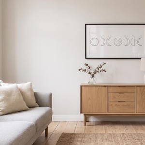Peut inclure: Une œuvre d'art murale rectangulaire dans un cadre noir présentant un dessin au trait noir des phases du cycle lunaire sur fond blanc, exposée au-dessus d'un buffet en bois clair avec trois tiroirs centraux et deux placards latéraux.