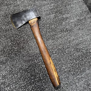 May include: A hand axe featuring a dark, weathered metal head with a sharp, curved blade edge and a smooth, straight wooden handle with a visible grain pattern. The metal head shows signs of use and age, securely attached to the wooden shaft.