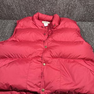 May include: A bright red puffer vest featuring a high collar, a front button closure with metallic snaps, and a visible L.L.Bean brand label sewn into the inner neckline. The garment has a quilted, padded construction with horizontal stitching.