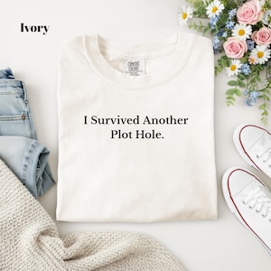 May include: An ivory-colored t-shirt with a crew neckline and the black text "I Survived Another Plot Hole." printed on the front. The shirt features a Comfort Colors brand label at the inner neck and is displayed with blue denim and white sneakers.