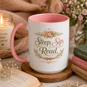 May include: A white ceramic mug with a pink interior and handle, featuring a floral wreath design surrounding the text "Steep, Sip, Read" in green and pink script. The mug is positioned on a round wooden coaster, displaying a clear, clean aesthetic.