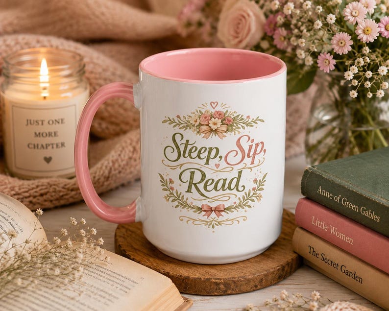 May include: A white ceramic mug with a pink interior and handle, featuring a floral wreath design surrounding the text "Steep, Sip, Read" in green and pink script. The mug is positioned on a round wooden coaster, displaying a clear, clean aesthetic.