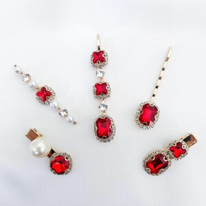 Puede incluir: Un conjunto de cinco accesorios para el cabello con monturas de metal dorado, cristales rojos rectangulares y cuadrados, y perlas blancas. El set incluye horquillas y pinzas adornadas con brillantes piedras de estrás transparentes.