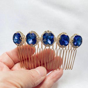 May include: A gold-toned hair comb featuring five large, round, deep blue faceted stones. Each stone is surrounded by a halo of small, clear, sparkling crystals. The comb has multiple thin, gold-coloured metal teeth for securing into hair.