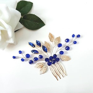 May include: A hair comb featuring gold-toned metal leaves and branches adorned with deep blue faceted beads and marquise-cut crystals. The design is arranged in a spray pattern, with the metal comb base integrated into the centre of the floral-inspired piece.