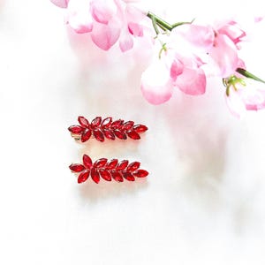 May include: Two hair clips featuring a leaf-shaped design composed of multiple marquise-cut red crystals set in a gold-toned metal frame. The hair accessories are arranged horizontally, showcasing a vibrant red colour and a faceted, reflective surface.