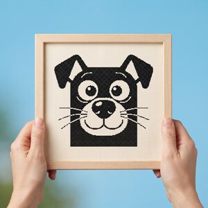 May include: A square cross-stitch embroidery featuring a stylized black and white dog face with large circular eyes, a rounded nose, and whiskers, framed in a light-colored wooden frame against a solid light blue background.