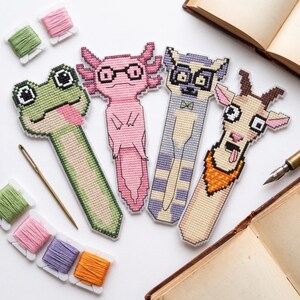 Peut inclure: Quatre marque-pages en point de croix avec des motifs d'animaux pixelisés : une grenouille verte tirant la langue, un axolotl rose à lunettes, un lémurien violet à lunettes et nœud papillon, et une chèvre beige avec un bandana orange.