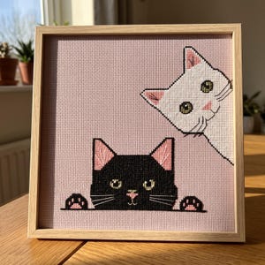 Peut inclure: Une broderie au point de croix carrée dans un cadre en bois clair représentant un chat noir qui dépasse du bas et un chat blanc qui regarde depuis le coin supérieur droit sur un fond rose uni. Les deux chats ont des oreilles et un nez roses.