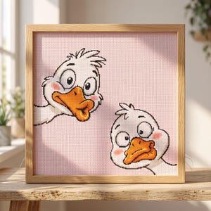 Peut inclure: Une broderie au point de croix carrée encadrée en bois clair, représentant deux oies blanches de dessin animé avec des becs orange et des joues roses sur un fond à grille rose pâle. Les oies ont de grands yeux expressifs et sont placées à différentes hauteurs.