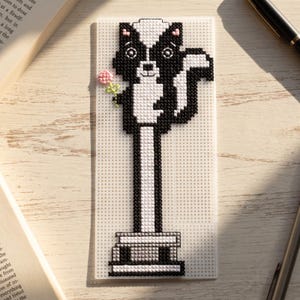Peut inclure: Un marque-page rectangulaire en plastique transparent avec un motif pixelisé au point de croix représentant une mouffette noire et blanche debout sur une pile de livres. La mouffette tient une petite fleur rose à tige verte.