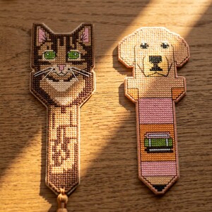 Peut inclure: Deux marque-pages en point de croix. Celui de gauche représente un chat tigré marron aux yeux verts avec un poitrail blanc. Celui de droite montre une tête de chien beige sur un corps en forme de crayon avec une gomme rose et un livre vert.