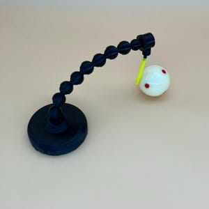 May include: A black articulated arm stand with a circular base, featuring a flexible ball-and-socket design. A bright neon yellow rod extends from the end, holding a smooth white sphere decorated with small red circular markings on its surface.