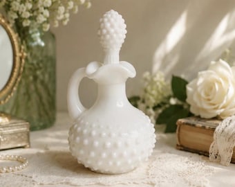 May include: A white milk glass decanter featuring a hobnail texture, a rounded body, a curved handle, and a ruffled rim. The decanter includes a matching removable stopper with a textured, pointed top, creating a cohesive and solid white appearance.