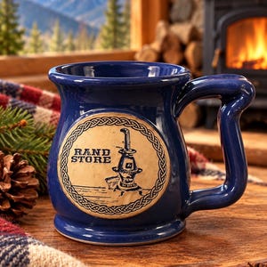 May include: A deep blue ceramic mug featuring a circular beige medallion on the front. The medallion displays a black illustration of a vintage potbelly stove and the text RAND STORE, surrounded by a decorative braided border pattern.