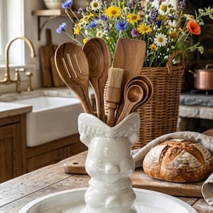 May include: A white ceramic utensil holder shaped like a stylized animal, containing a collection of wooden kitchen tools including a slotted spoon, a solid spoon, a flat spatula, a pastry brush, and several measuring spoons on a white tray.