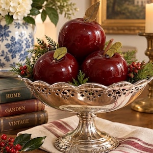 May include: A silver-toned pedestal bowl with a scalloped, pierced rim containing three glossy, deep red decorative apples. The apples are accented with small green leaves and stems, nestled among sprigs of evergreen foliage and tiny red berries.