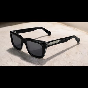 Può includere: Occhiali da sole neri con montatura rettangolare spessa e lenti scure. Le aste presentano un dettaglio metallico color argento vicino alla cerniera e la montatura ha una finitura lucida con un bordo inferiore leggermente traslucido.