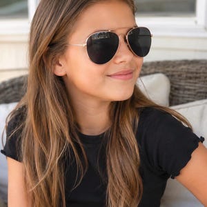 Peut inclure: Lunettes de soleil aviateur avec de fines montures en métal or rose et des verres noirs opaques. Ces lunettes présentent une forme en goutte d'eau classique avec un double pont sur le dessus, offrant un style épuré et une protection solaire.