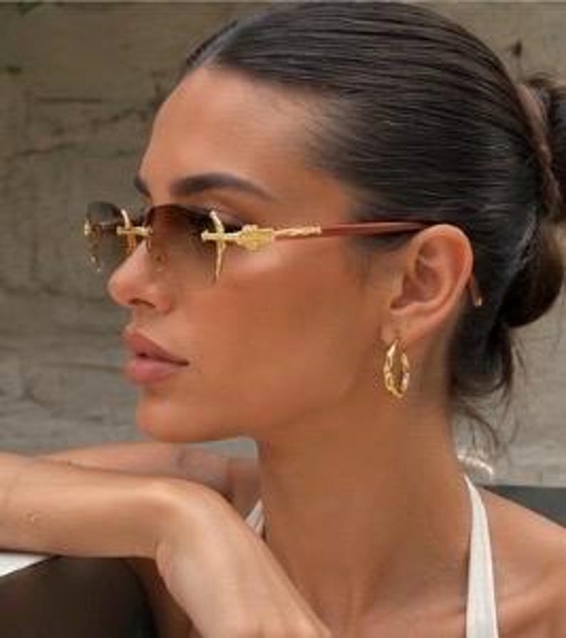 May include: Rimless sunglasses featuring gradient brown lenses and gold-tone metal temples with intricate detailing. The bridge and hinges display ornate gold-colored accents, and the thin arms transition into a dark brown material at the tips.